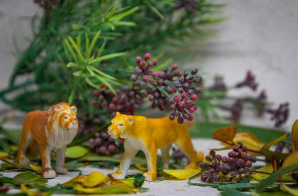 Close-up of miniature lion and lioness figurines surrounded by lush green foliage and flowers.