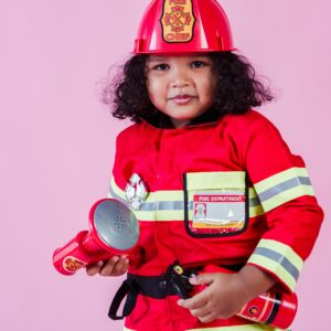 Little ethnic child wearing fireman costume with fire extinguisher and loudspeaker toy with hardhat and standing on pink background and looking at camera