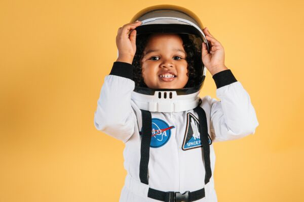 Smiling little African American girl in white space suit looking at camera touching helmet on orange background