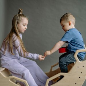 Two siblings share a playful moment on a wooden seesaw indoors.